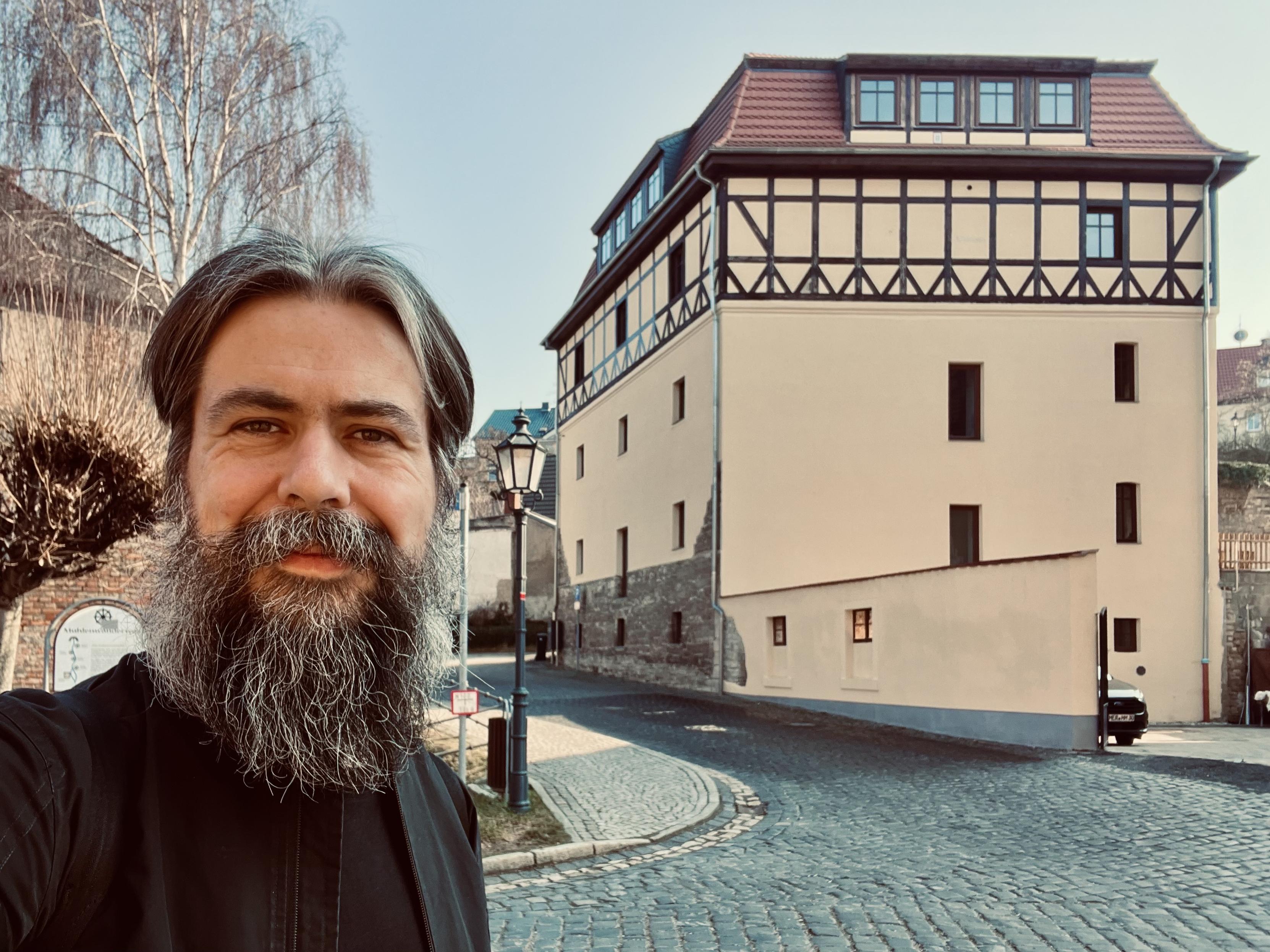 Selfie eines bärtigen Mannes mit langen Haaren vor einer frisch sanierten historischen Mühle mit Fachwerk im oberen Stockwerk. Das Gebäude steht an einer gepflasterten Straße mit Laterne und Bäumen, bei klarem Himmel und Tageslicht.